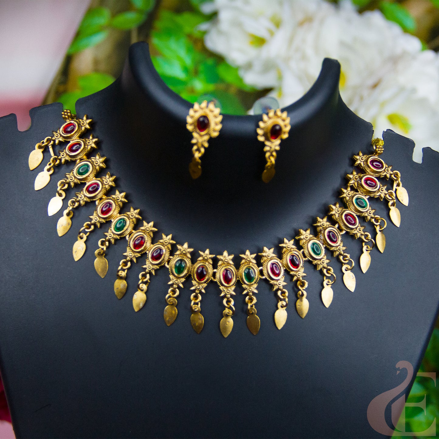 Gold-toned jewelry set displayed on a black mannequin bust, featuring a necklace and matching earrings with red and green stone accents.