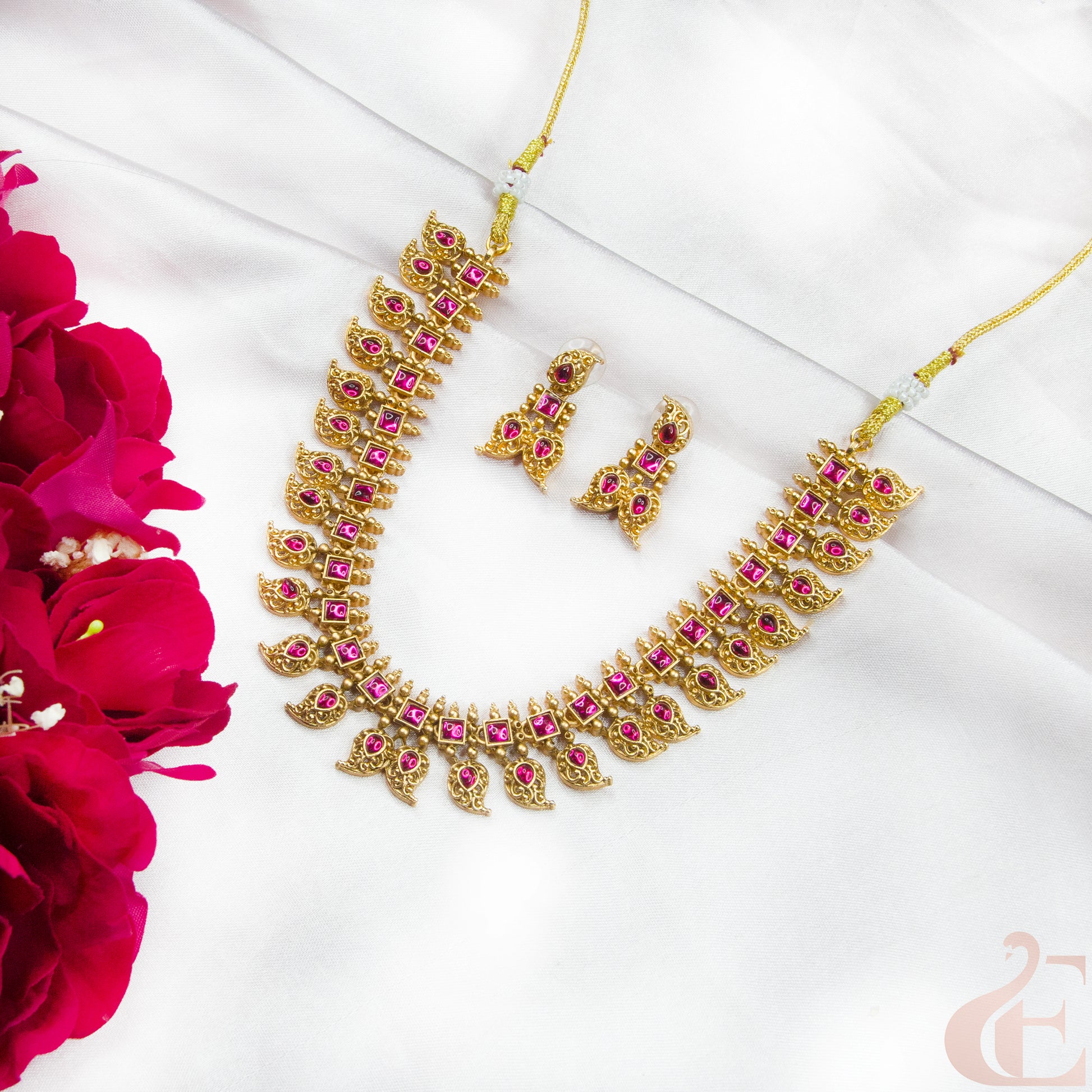 Gold necklace with pink stones and matching earrings on a white surface with red flowers.
