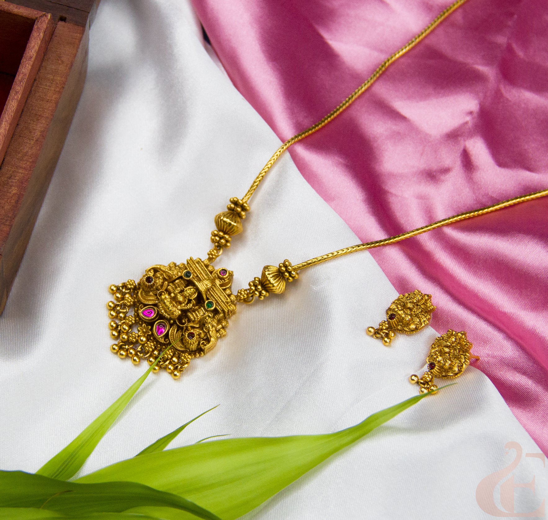 Gold necklace and earrings on a white and pink fabric background with green leaves.