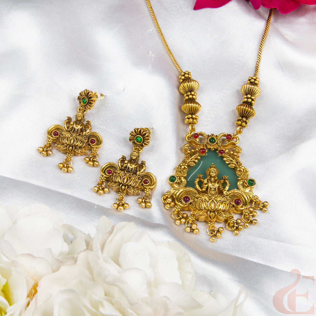 Gold-toned jewelry set featuring a necklace and matching earrings with ornate filigree detailing, green stone accents, and red gemstones, arranged on white satin fabric with white flowers.