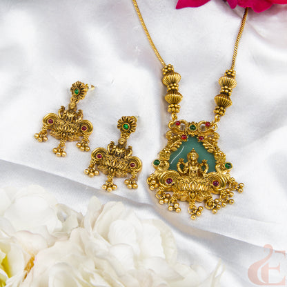 Gold-toned jewelry set featuring a necklace and matching earrings with ornate filigree detailing, green stone accents, and red gemstones, arranged on white satin fabric with white flowers.