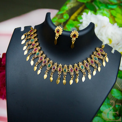 Gold-toned traditional necklace and matching drop earrings displayed on a black jewelry bust. The necklace features oval red and green stone accents with dangling gold leaf-shaped charms, set against a softly blurred floral background.