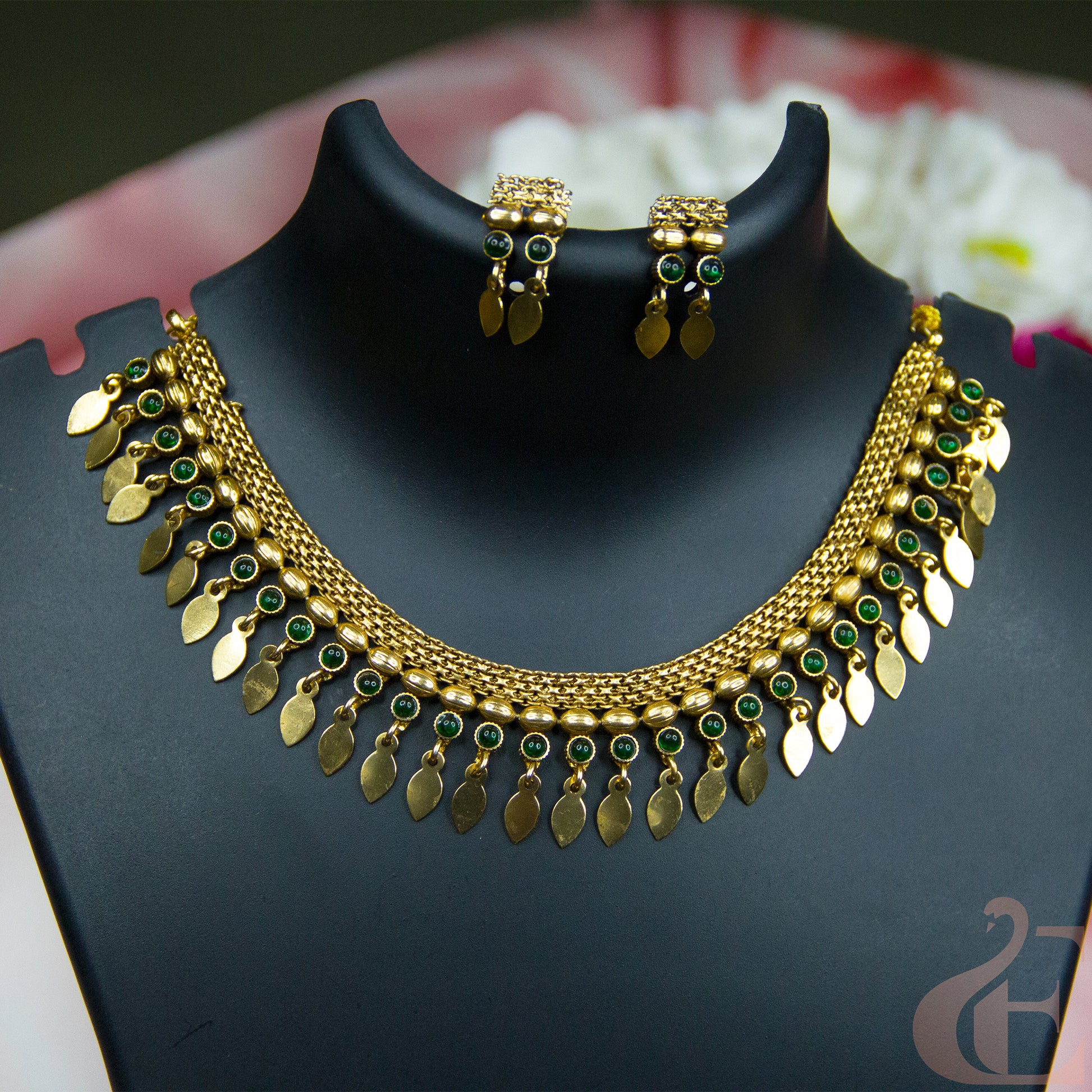 Gold-tone Indian style necklace and matching earrings set displayed on a black jewelry bust. The necklace features a textured chain with dangling gold leaf-shaped charms and small round green stone accents.