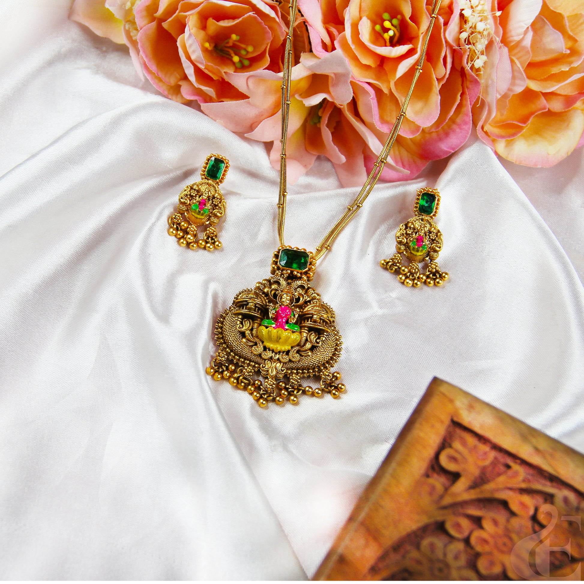 Gold necklace with green and red stones on a white fabric background with pink flowers.