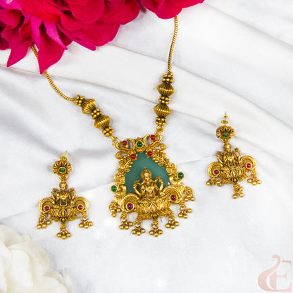 Gold-toned traditional Indian jewelry set featuring a necklace and matching earrings with a central green stone inlay and red and green stone accents, arranged on a white fabric background with pink flowers at the top.