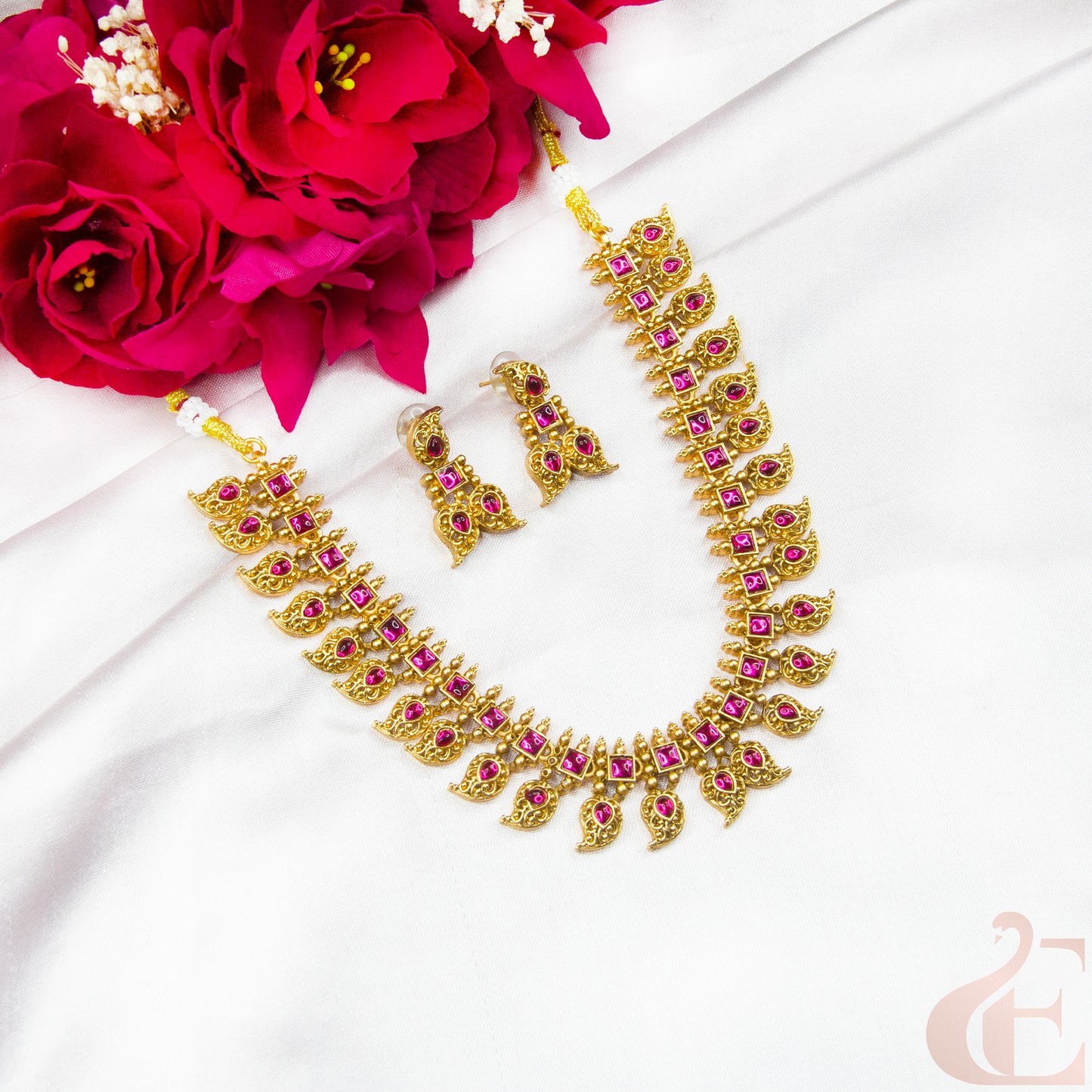 Gold necklace with pink stones and matching earrings on a white surface with red flowers.