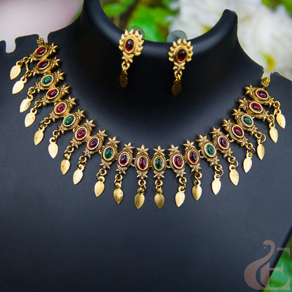 Gold-plated Indian jewelry set featuring a statement collar necklace and matching drop earrings, embellished with red and green stones on a black display bust.