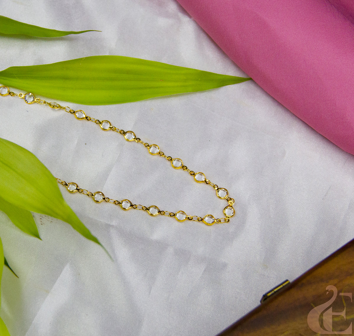 Gold chain necklace on a white fabric background with green leaves and a pink fabric.