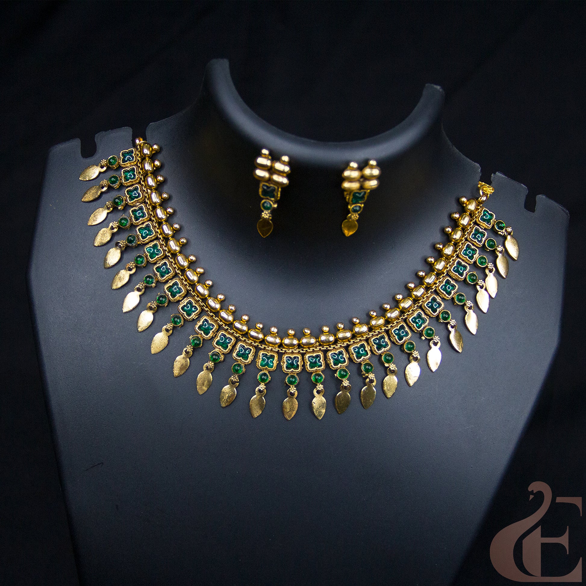 Gold-plated statement necklace and matching earrings with green stone accents, displayed on a black jewelry bust against a dark background.