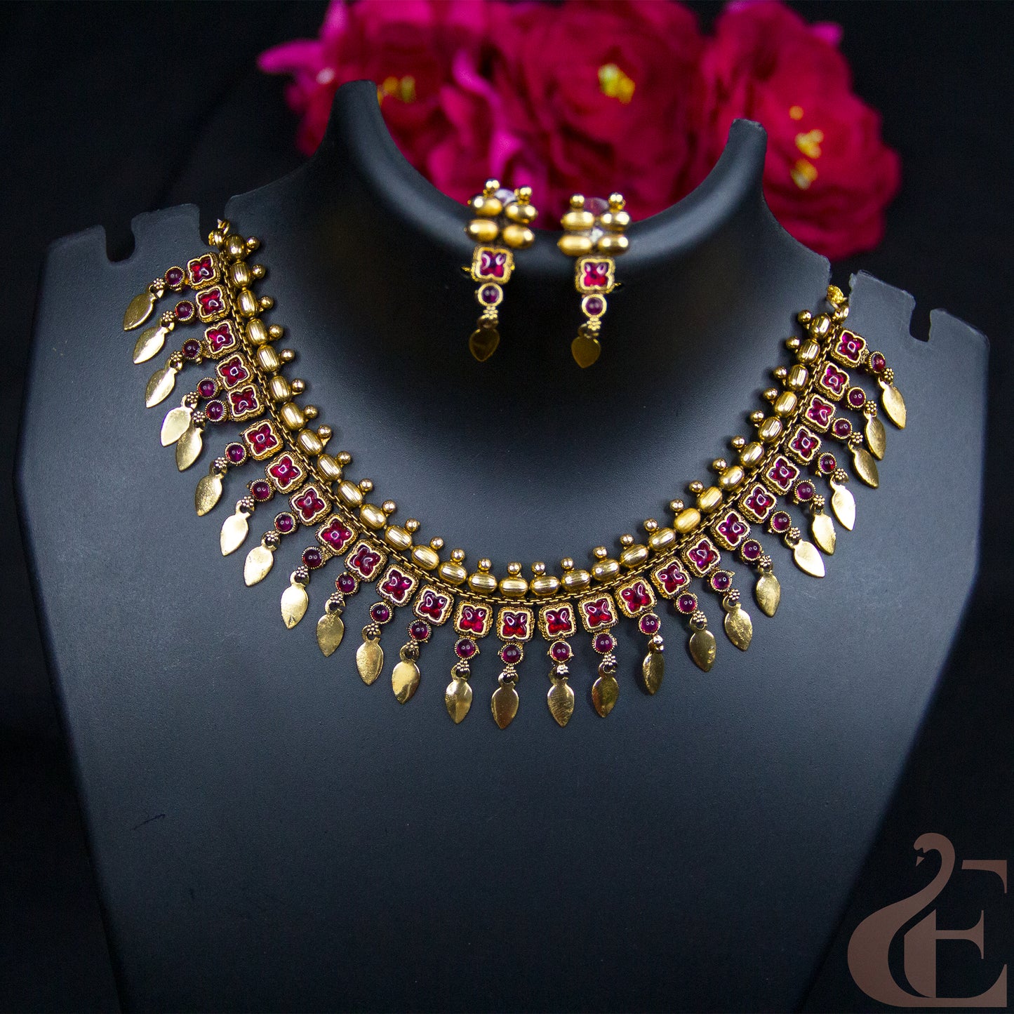 Gold-toned Indian-style jewelry set featuring a necklace and matching earrings displayed on a black mannequin bust, with dark pink flowers in the background.