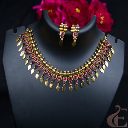 Gold-toned Indian-style jewelry set featuring a necklace and matching earrings displayed on a black mannequin bust, with dark pink flowers in the background.