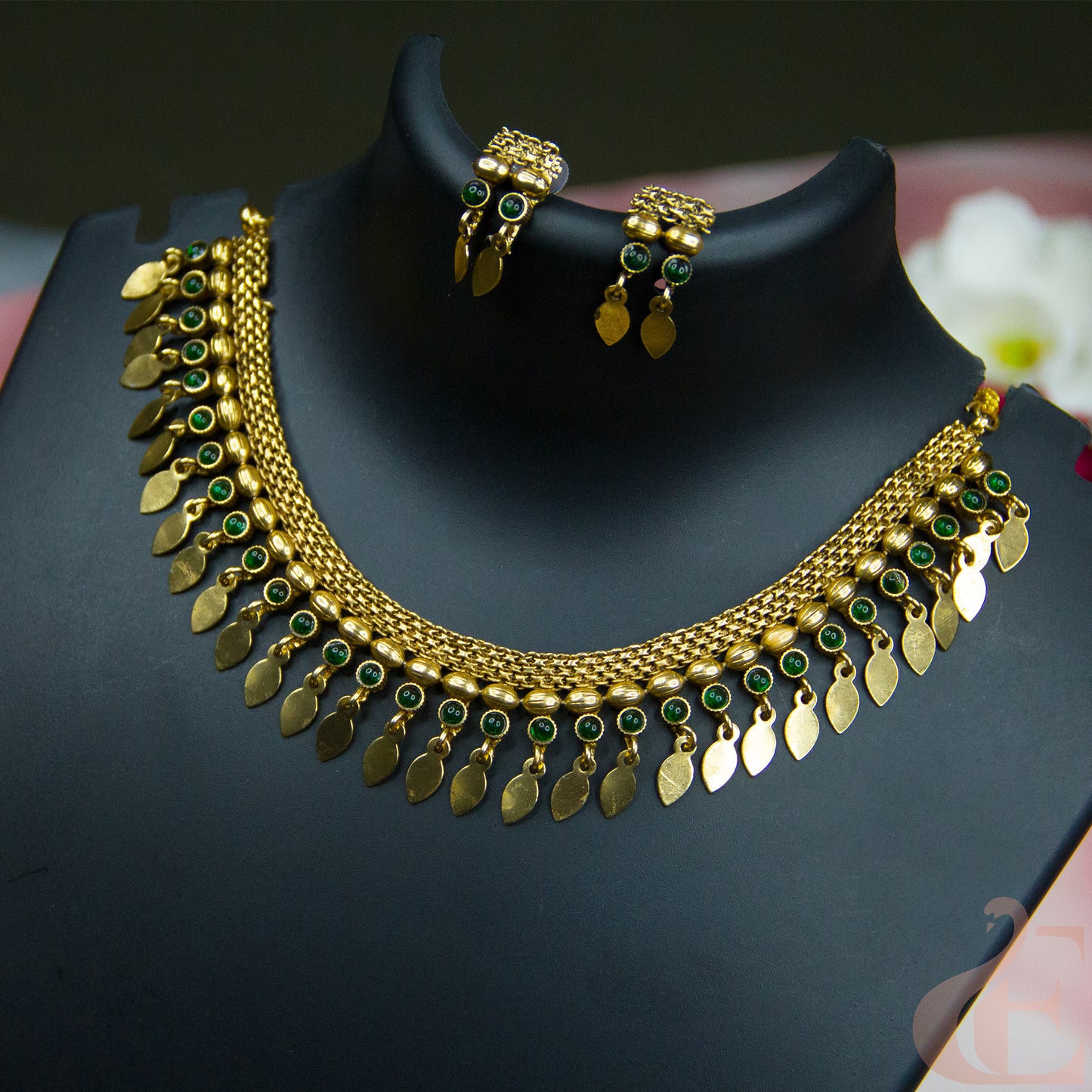 Gold-toned traditional necklace and matching earrings displayed on a black jewelry bust. The necklace features multiple rows of gold leaf-shaped charms, small green stone accents, and a beaded chain.