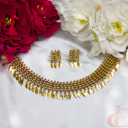Gold-toned traditional jewelry set featuring a multi-strand necklace with dangling teardrop-shaped charms and matching drop earrings, arranged on a white fabric background with red and white flowers.