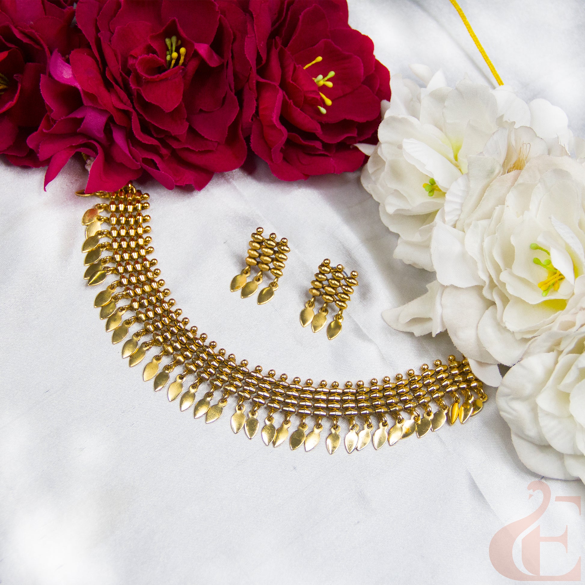 Gold-toned traditional necklace and matching earrings displayed on a white fabric surface, styled with red and white flowers around the edges.