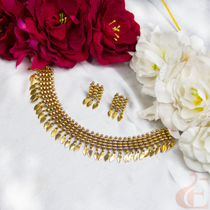 Gold-toned traditional necklace and matching earrings displayed on a white fabric surface, styled with red and white flowers around the edges.
