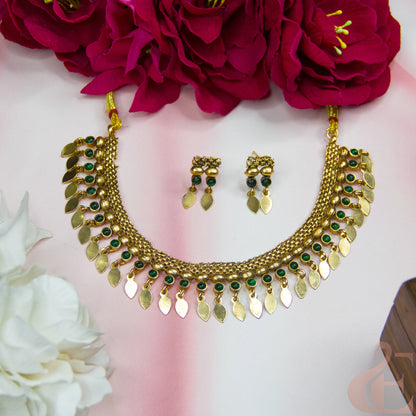 Gold-toned traditional necklace and matching earrings with green stone accents and dangling leaf-shaped charms, arranged on a pale pink background with red and white flowers as styling props.
