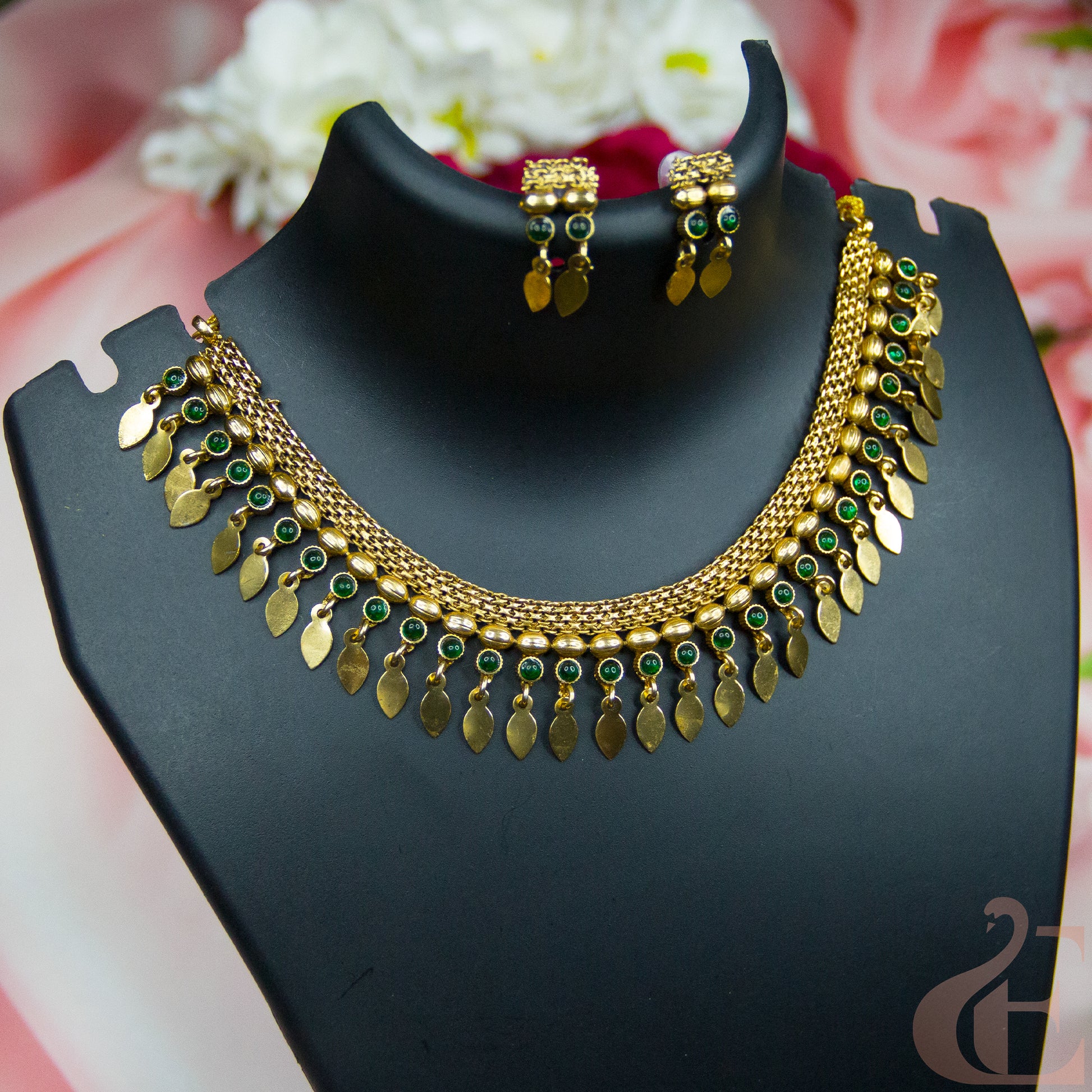 Gold-toned traditional necklace and matching earrings displayed on a black jewelry bust, with green stone accents and dangling leaf-shaped charms, against a soft pink floral background.