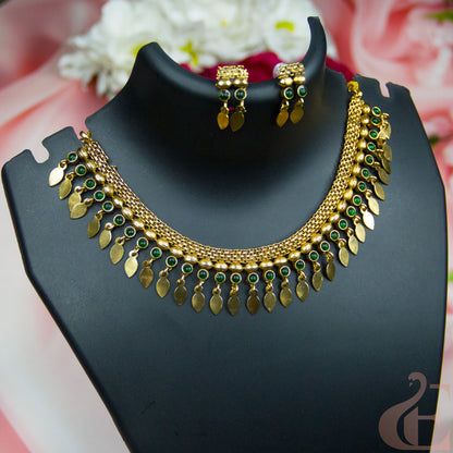 Gold-toned traditional necklace and matching earrings displayed on a black jewelry bust, with green stone accents and dangling leaf-shaped charms, against a soft pink floral background.