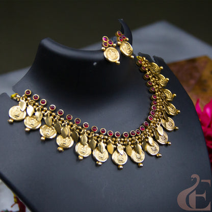 Gold coin pendant necklace and matching earrings displayed on a black jewelry bust. The necklace features multiple gold coin pendants, small gold beads, and rows of red gemstone accents, with matching coin-style earrings hanging above the bust.