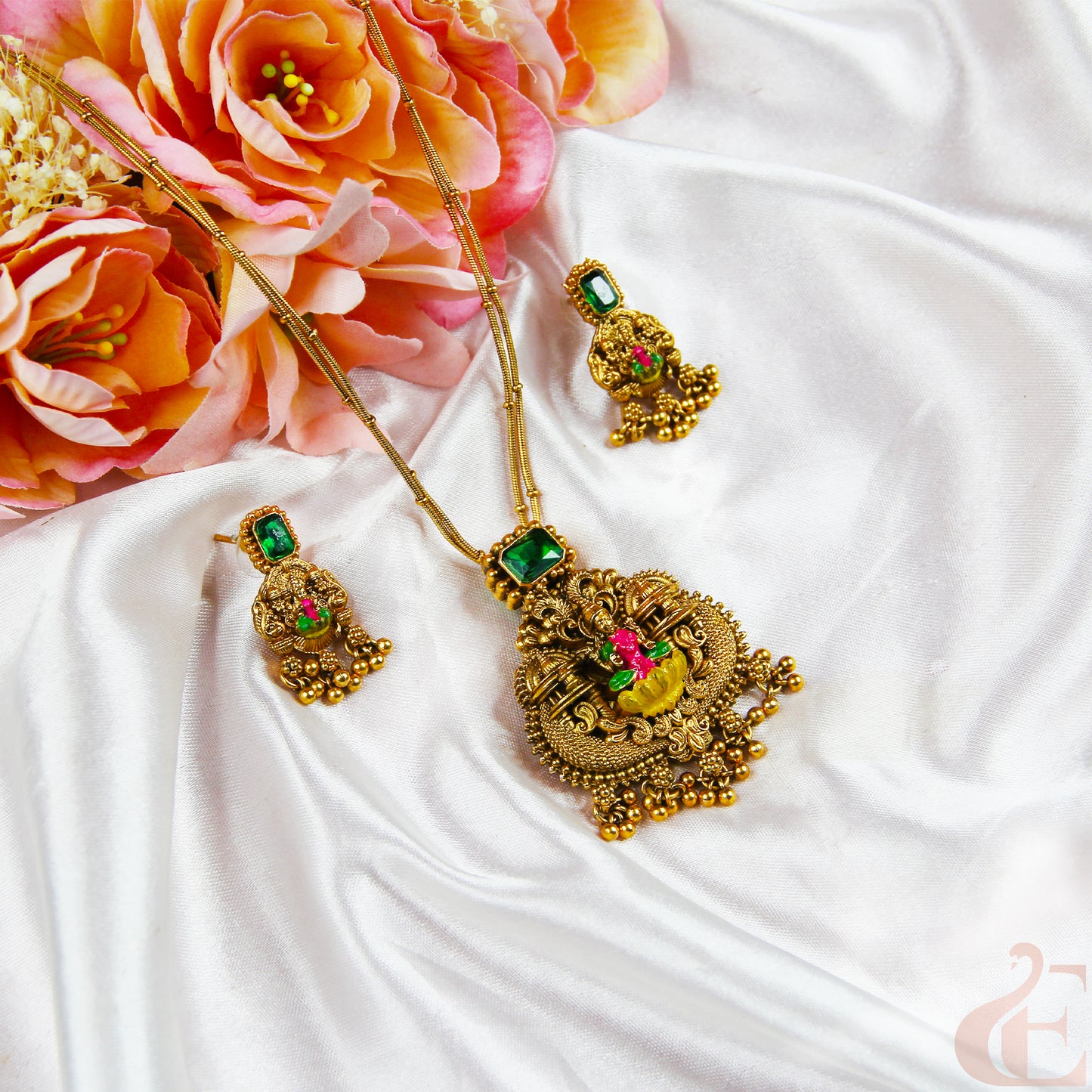Gold necklace with green and red gemstones on a white fabric background with pink flowers.