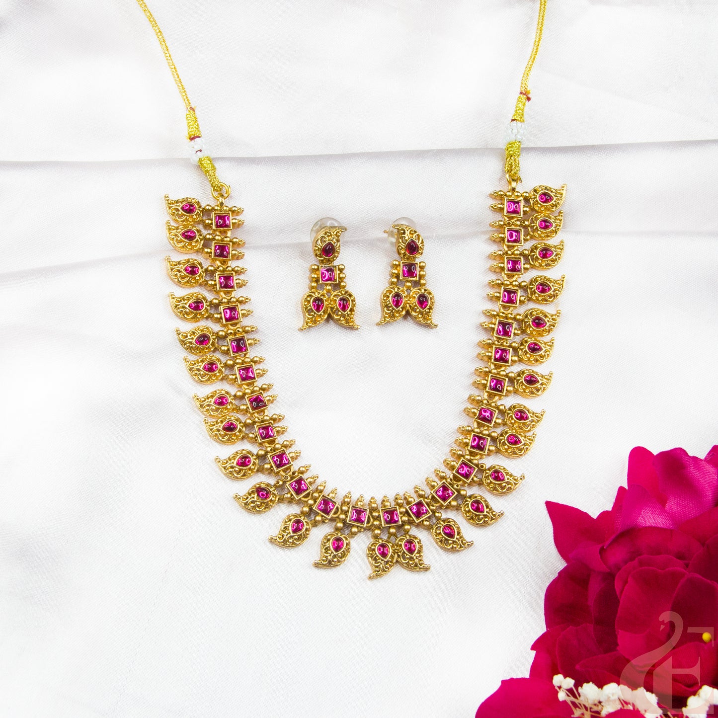 Gold necklace with pink stones and matching earrings on a white background with a red flower.