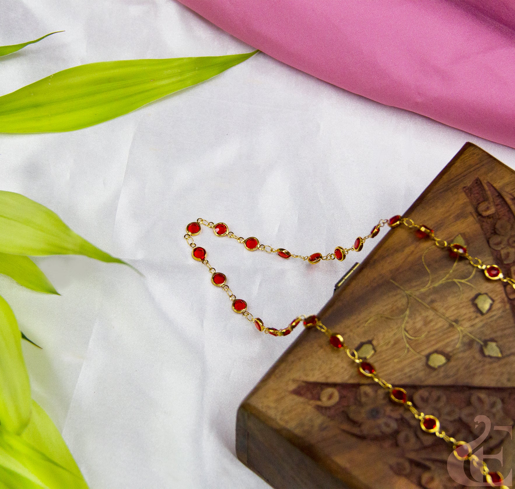 Gold and red beaded necklace on a wooden box with pink fabric and green leaves in the background