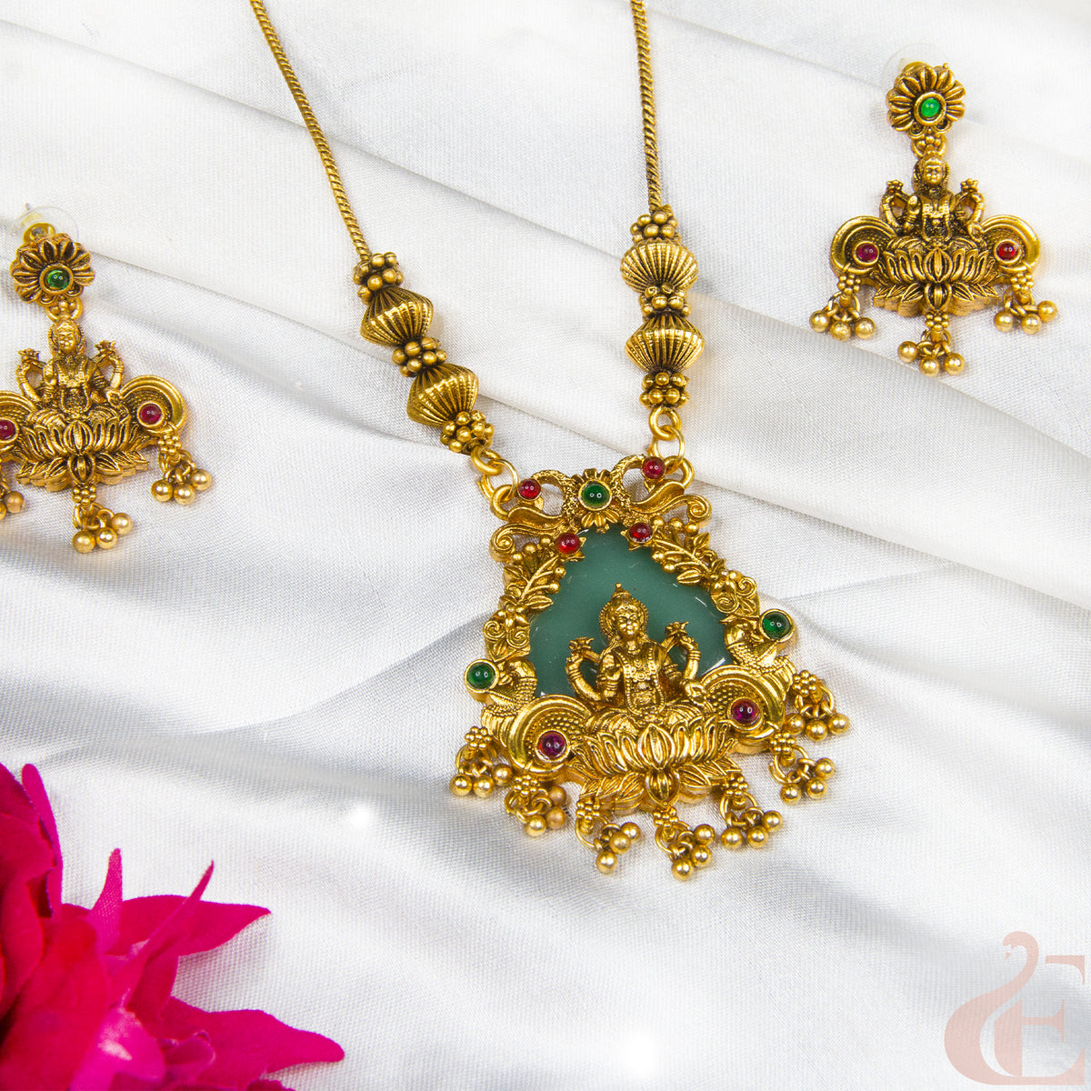 Gold-toned traditional jewelry set featuring a detailed pendant necklace and matching earrings with a central green stone and small red and green stone accents, displayed on white draped fabric with a pink flower in the corner.