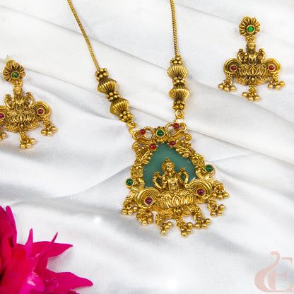 Gold-toned traditional jewelry set featuring a detailed pendant necklace and matching earrings with a central green stone and small red and green stone accents, displayed on white draped fabric with a pink flower in the corner.
