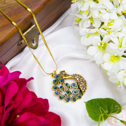 Gold necklace with a peacock pendant on a white fabric background with flowers and a wooden box.