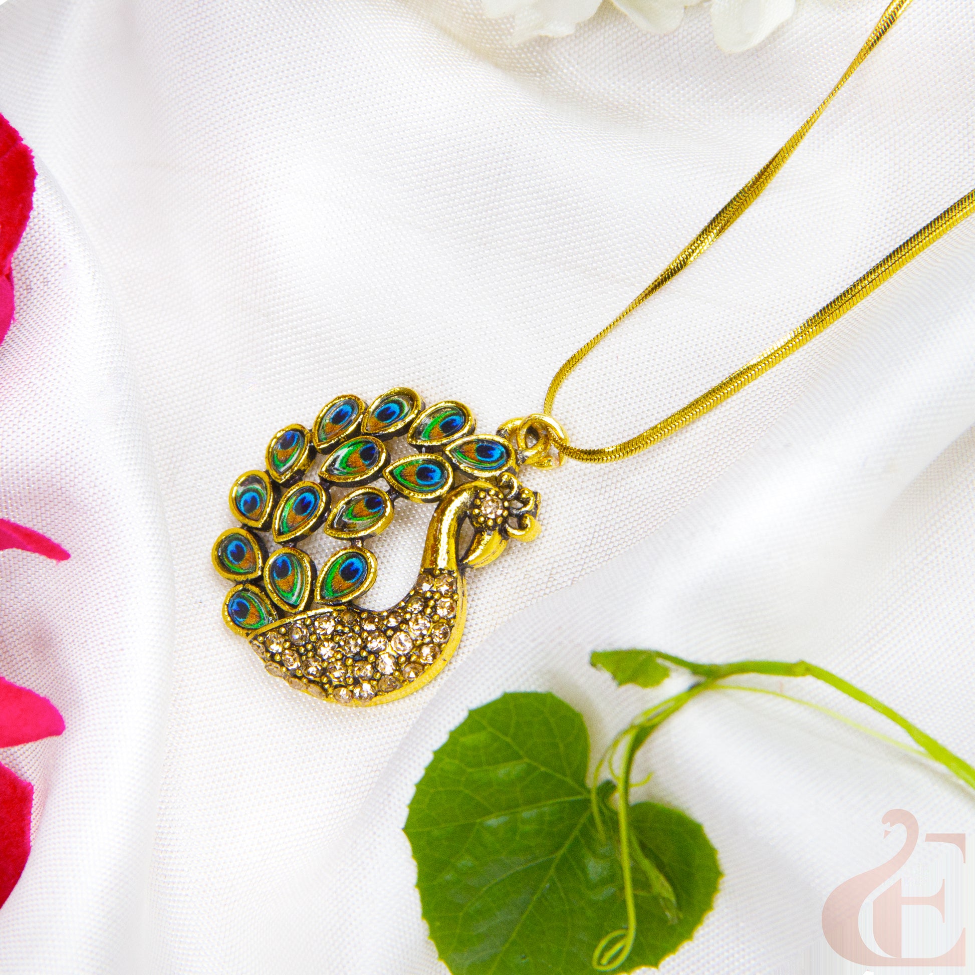 Gold peacock pendant necklace on a white fabric background with green leaves and pink flowers.