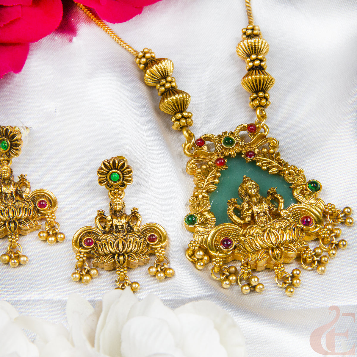 Gold-toned traditional Indian jewelry set featuring a pendant necklace and matching earrings with a green stone center, red and green gemstone accents, and ornate filigree detailing, arranged on white fabric with pink and white flowers.