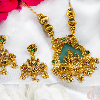 Gold-toned traditional Indian jewelry set featuring a pendant necklace and matching earrings with a green stone center, red and green gemstone accents, and ornate filigree detailing, arranged on white fabric with pink and white flowers.