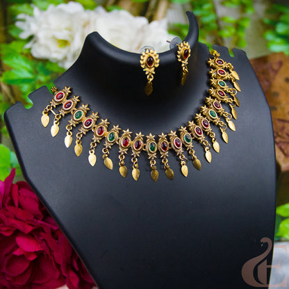 Gold-toned traditional Indian jewelry set featuring a necklace and matching earrings with red and green stone accents, displayed on a black jewelry bust with blurred flowers and foliage in the background.