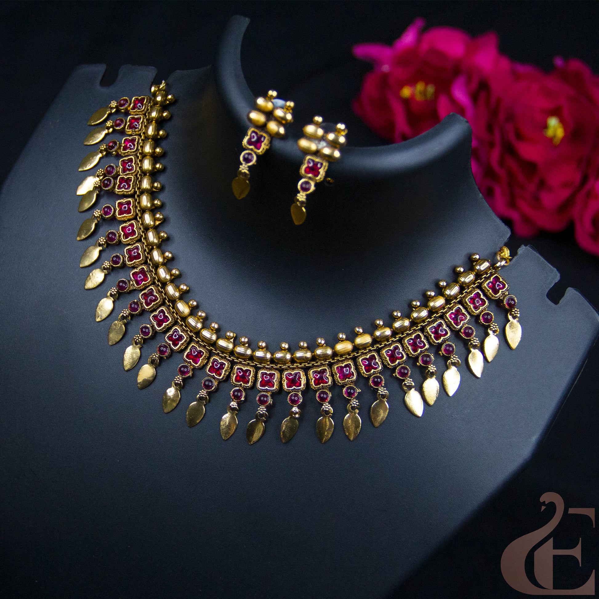 Gold-plated Indian style necklace and matching earrings with red square and round stones, displayed on a black jewelry bust with pink flowers in the background.