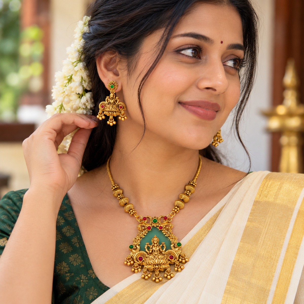 A woman wearing an ornate gold necklace and matching earrings with green and red stone accents, styled with a white and gold sari and white flowers in her hair.