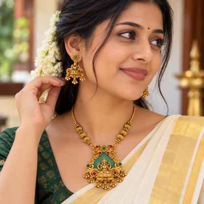 A woman wearing an ornate gold necklace and matching earrings with green and red stone accents, styled with a white and gold sari and white flowers in her hair.
