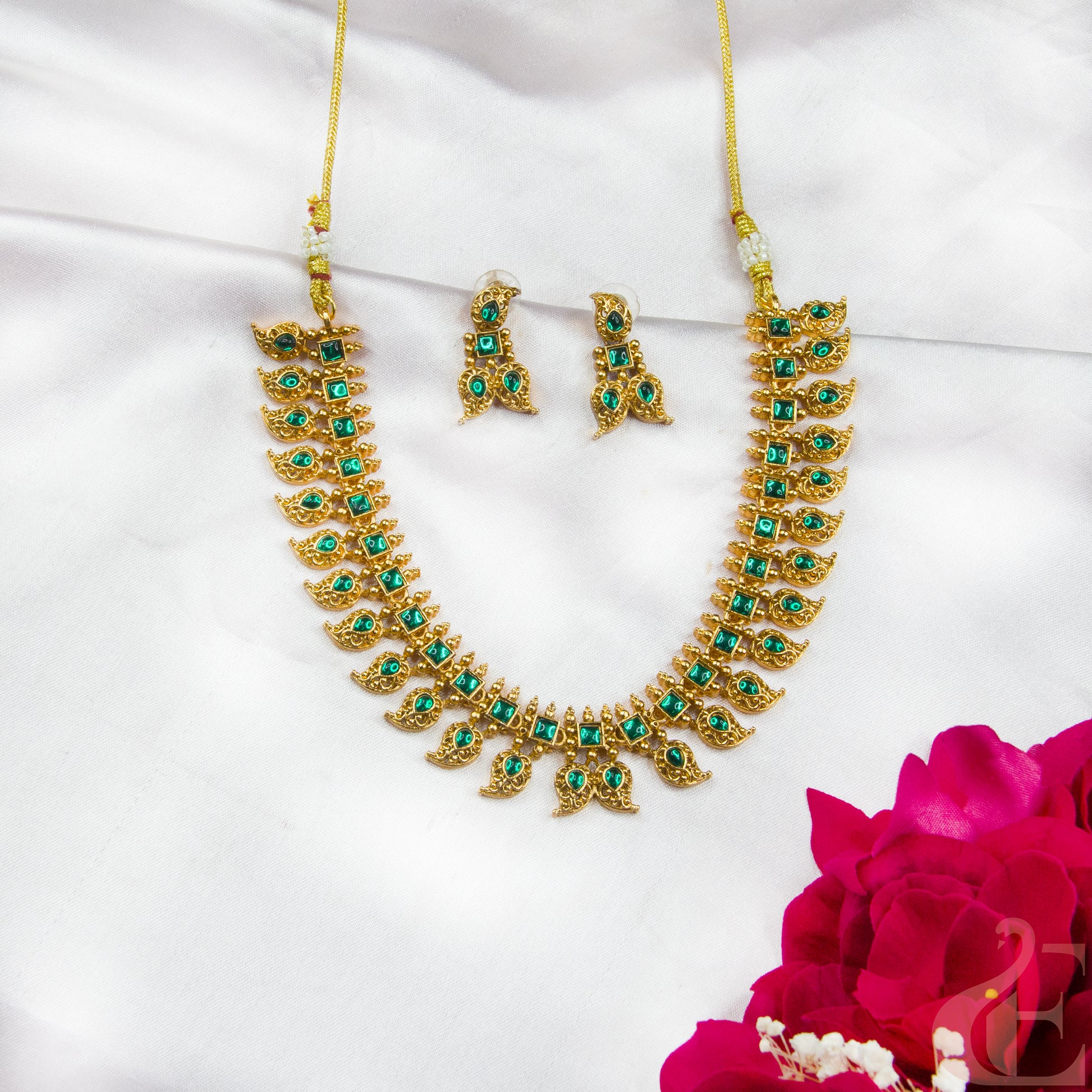 Gold necklace with green gemstones and matching earrings on a white background with a pink flower.