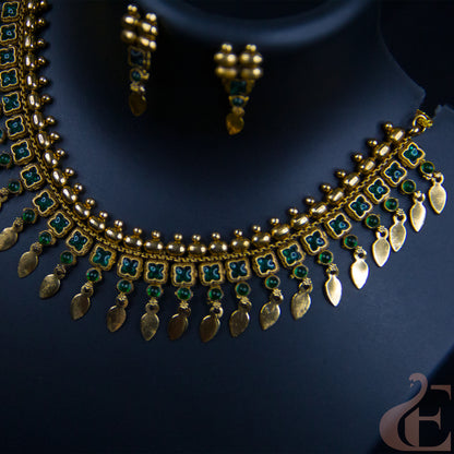 Gold-toned traditional necklace and matching earrings displayed on a dark navy jewelry bust. The necklace features green stone floral motifs and dangling gold leaf-shaped charms.